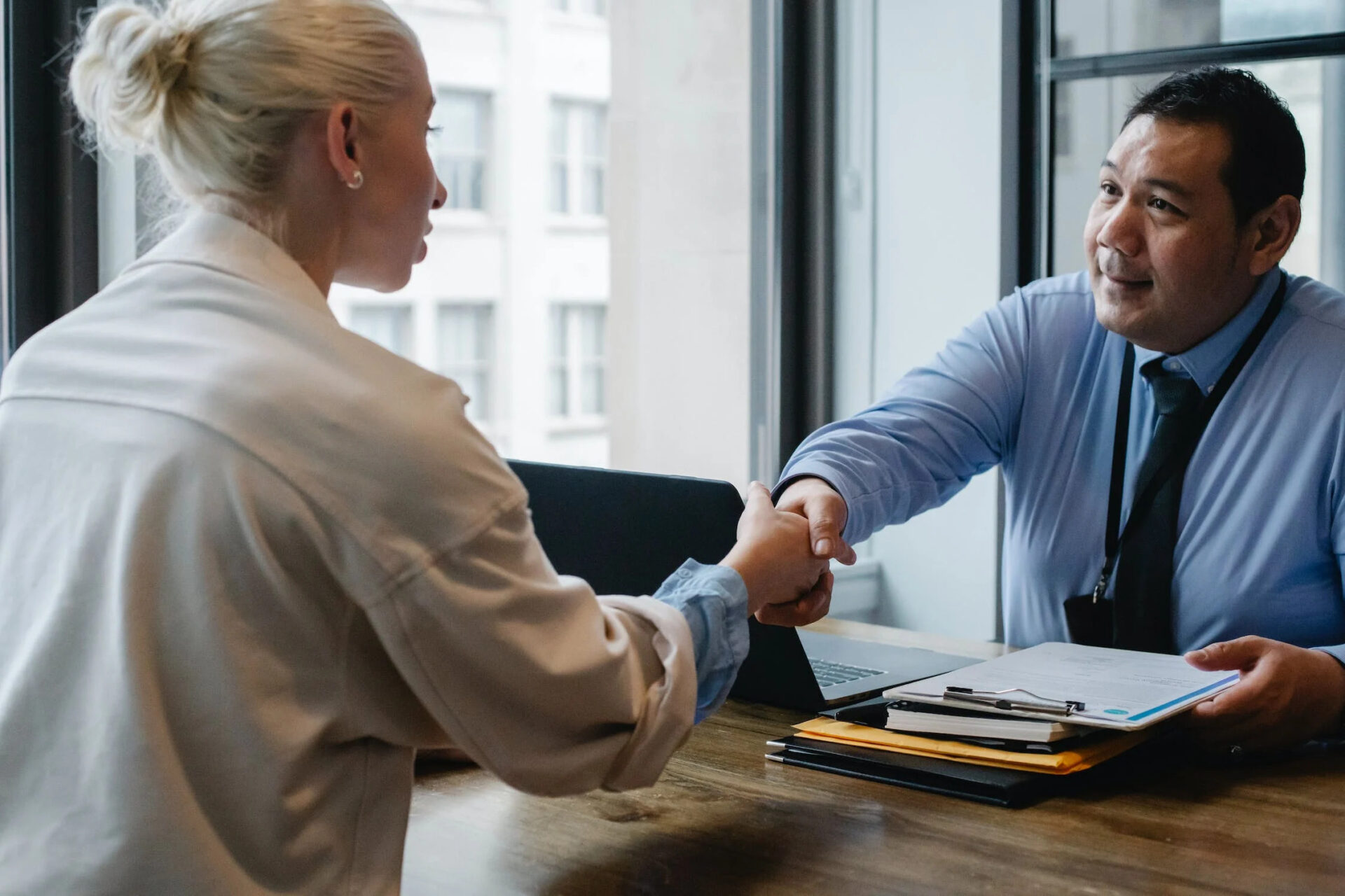A professional looking person wearing a blue shirt and tie is shaking hands with a blonde haired woman in an office over a stack of documents